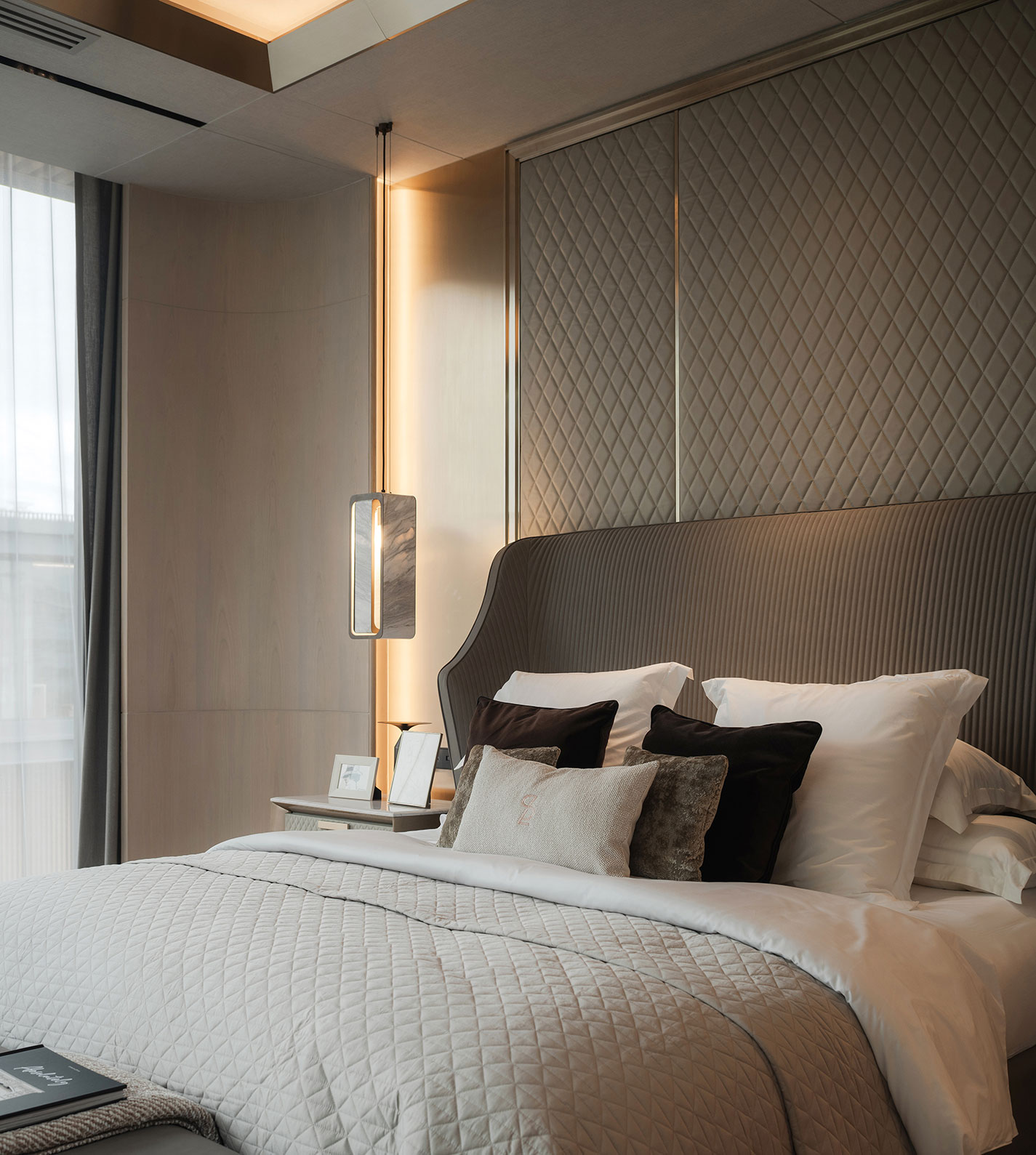 Luxurious bedroom with quilted beige headboard, layered pillows, and soft lighting. Elegant pendant light hangs beside the neatly arranged bed.