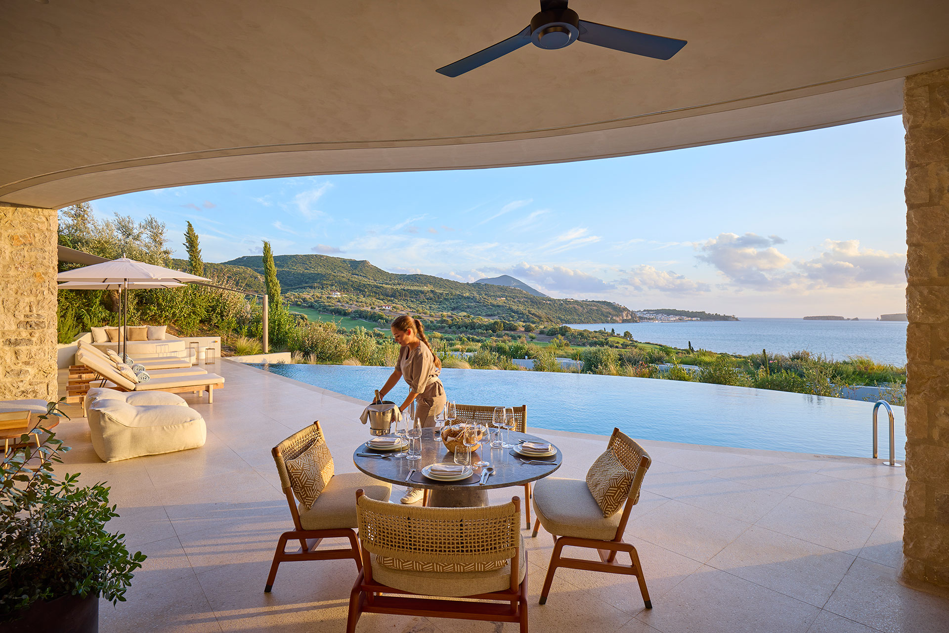Outdoor terrace overlooking a serene infinity pool and ocean. A woman sets a dining table with elegant place settings, conveying tranquility and luxury.