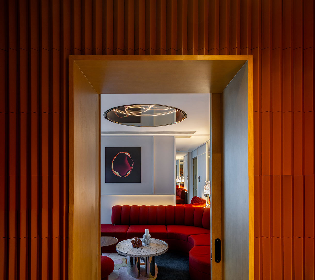 Modern interior with a warm, orange-hued entryway leading to a stylish living room. Features a red curved sofa, a round table, and abstract wall art.