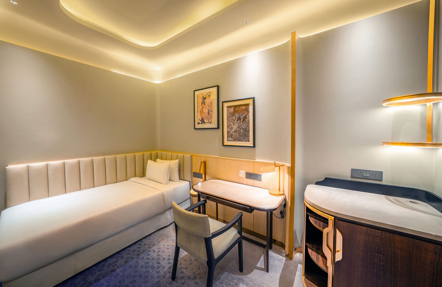 Modern lounge room with single bed, beige upholstered headboard, desk, chair, art on walls, soft lighting, creating a cozy, minimalist ambiance.