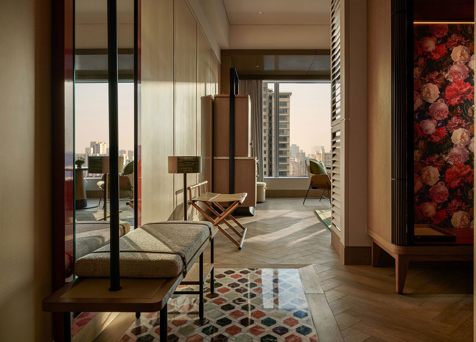 Chic hotel room with modern decor, wooden floors, geometric rug, and floral wallpaper. Large window reveals an urban skyline at sunset. Cozy and elegant ambiance.