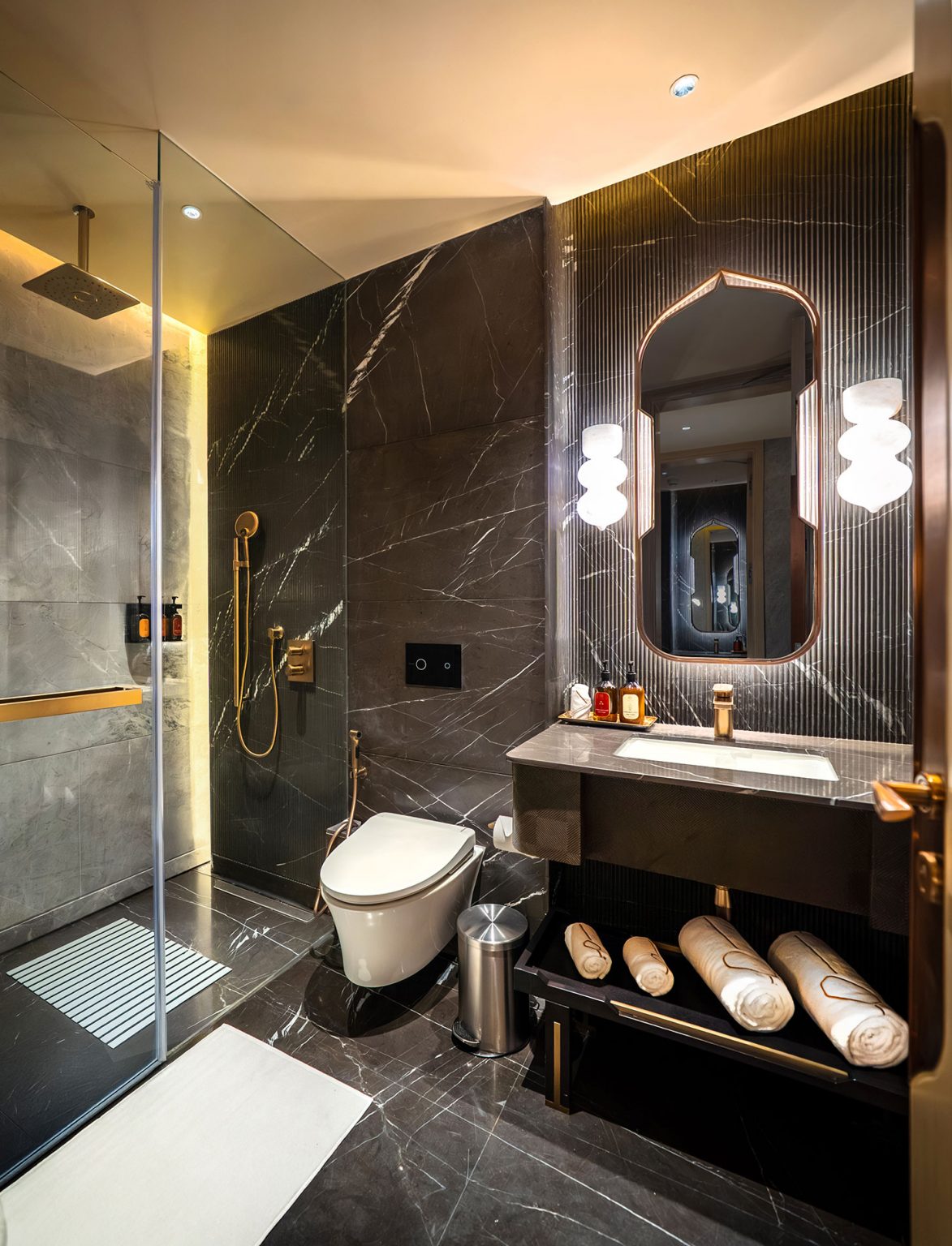 Luxurious bathroom with dark marble walls and floor, elegant rainfall shower, backlit mirror, sleek sink, and neatly stacked towels, exuding modern elegance.