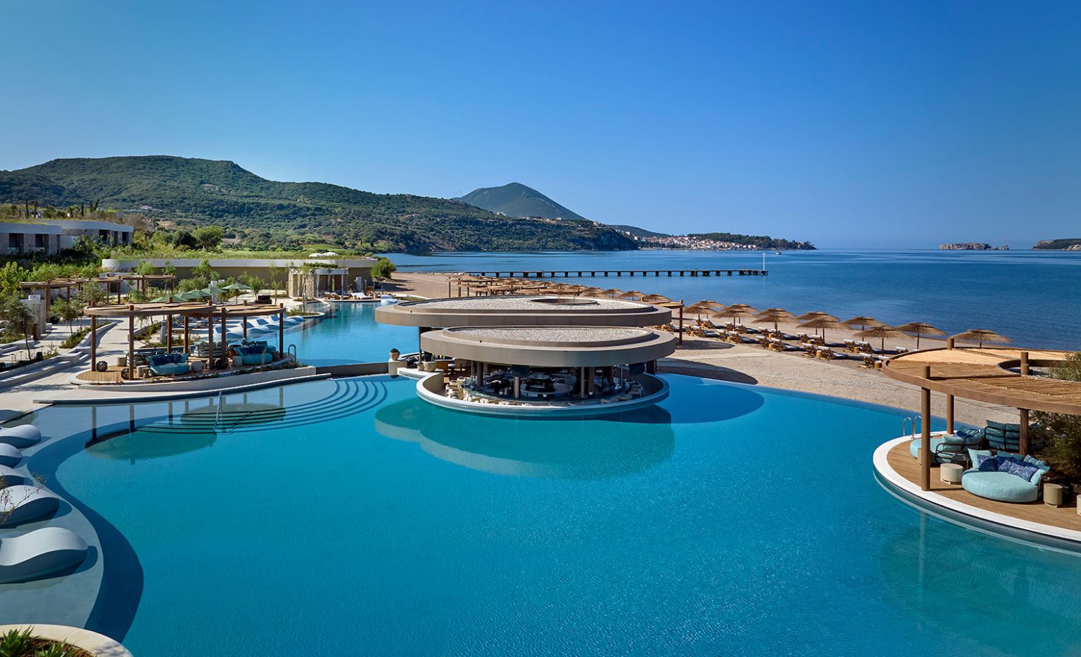 Expansive luxury resort infinity pool featuring a unique circular swim-up bar, surrounded by lounge areas, a sandy beach with umbrellas, and a calm ocean view with rolling green hills in the background.