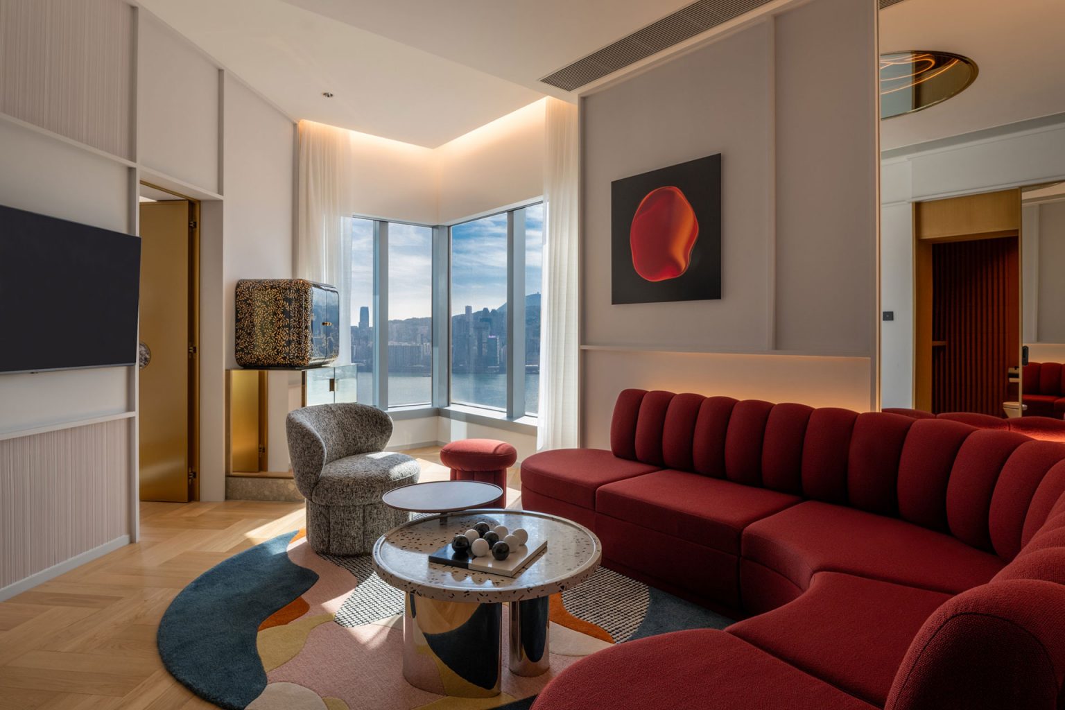 Modern living room with a red sectional sofa, abstract wall art, and a round coffee table. Large windows offer a city skyline view, creating a cozy yet sophisticated atmosphere.