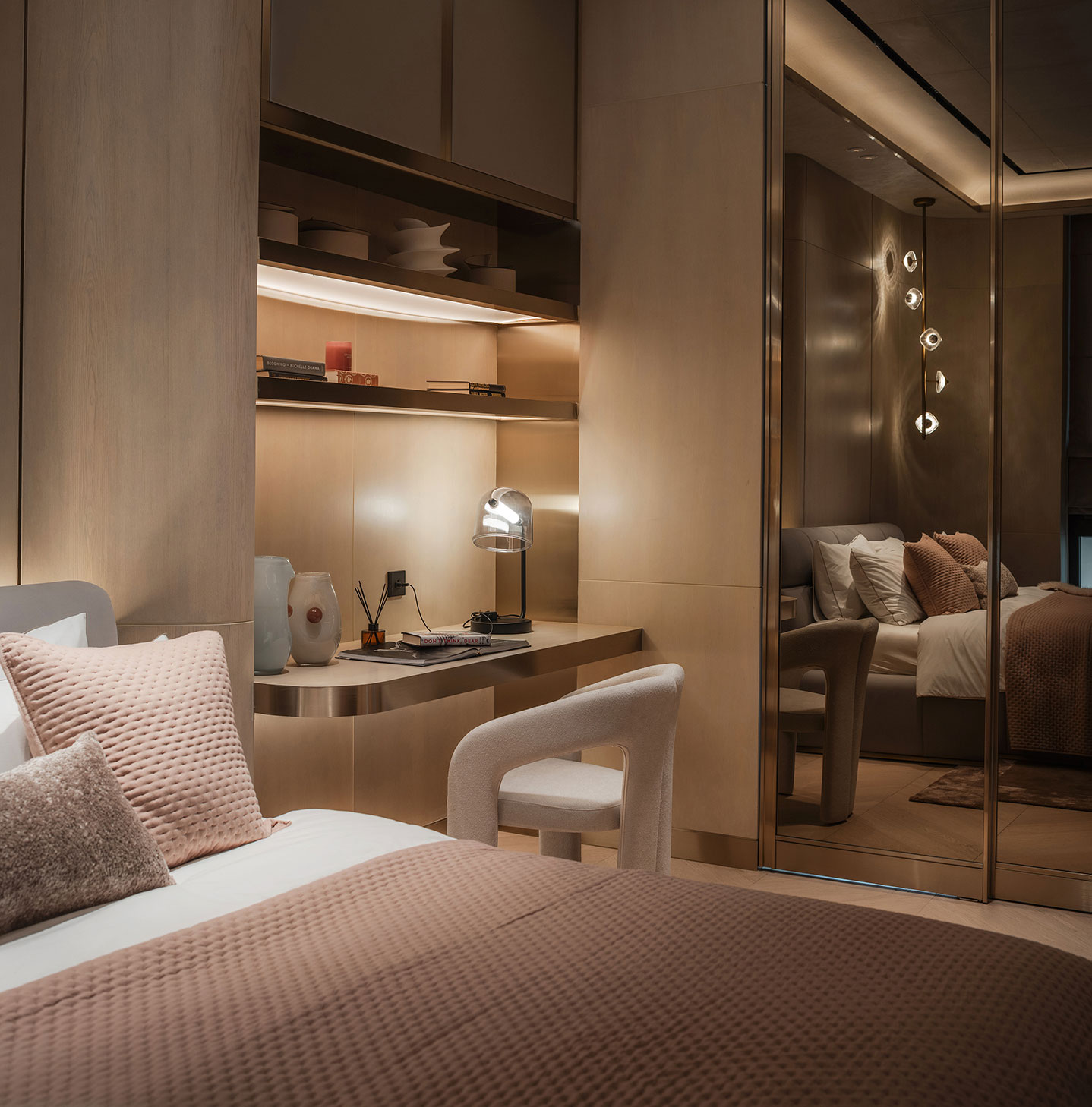Cozy bedroom with warm lighting and neutral tones. Features a cushioned bed, wooden desk, modern lamp, and reflective mirrored closet doors.