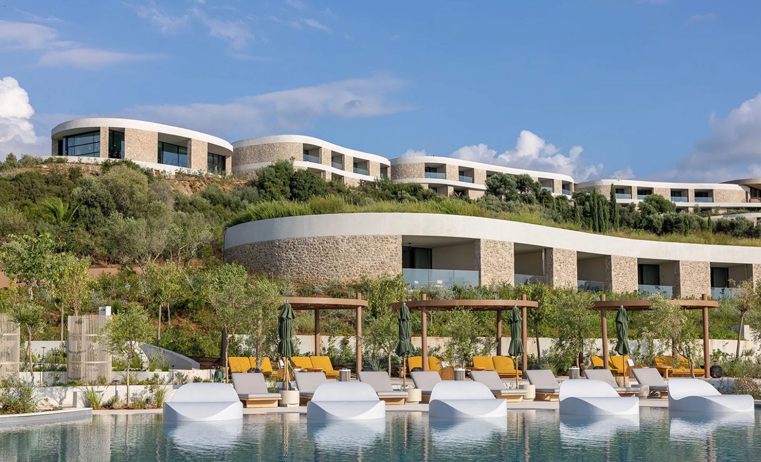 Luxury resort pool with white in-water loungers in the foreground, leading to terraced, modern stone buildings with green roofs built into a lush, green hillside under a partly cloudy sky.