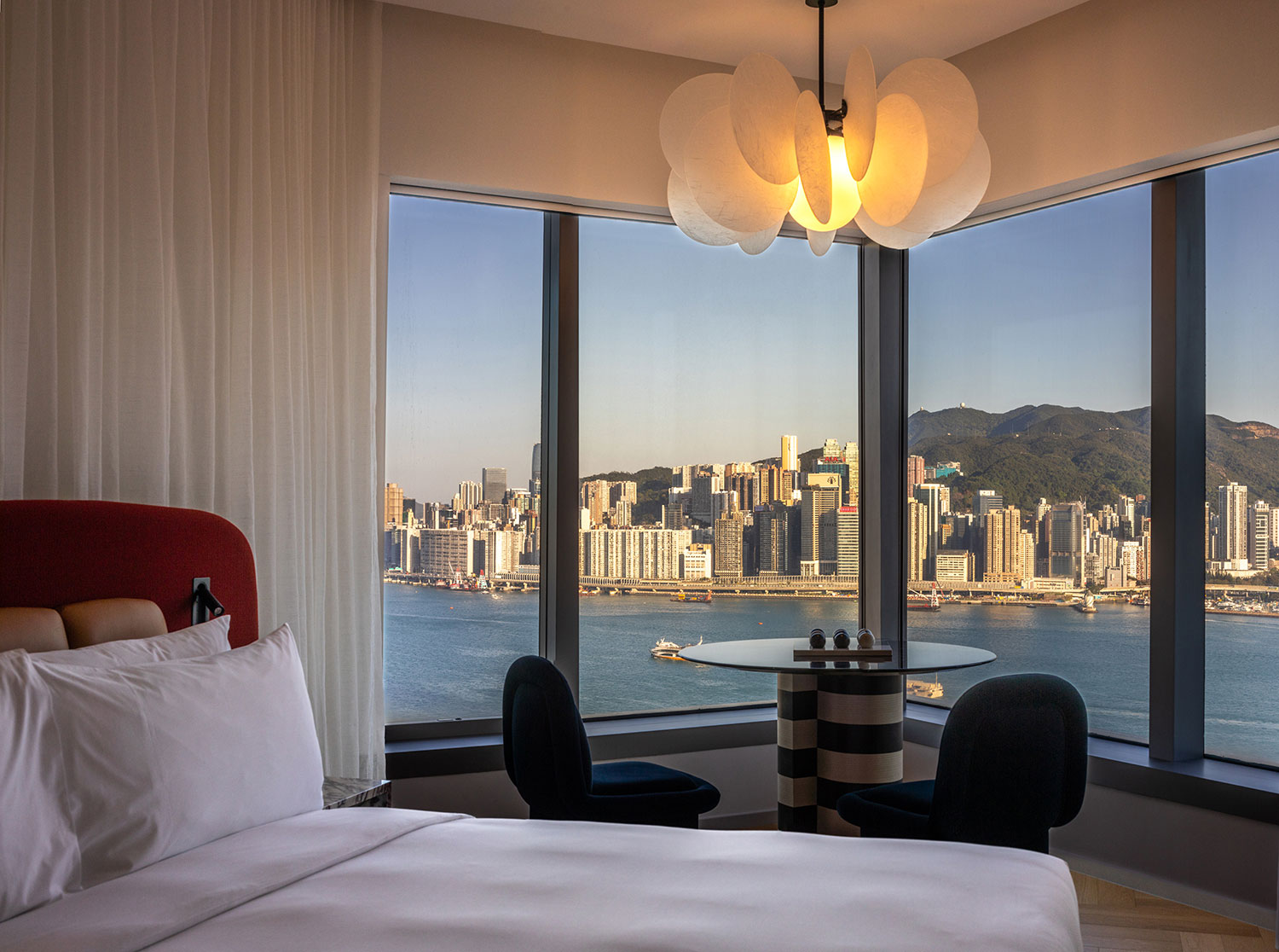 Hotel room with modern decor overlooking a city skyline and harbor. Large windows offer a panoramic view. Soft lighting creates a cozy atmosphere.