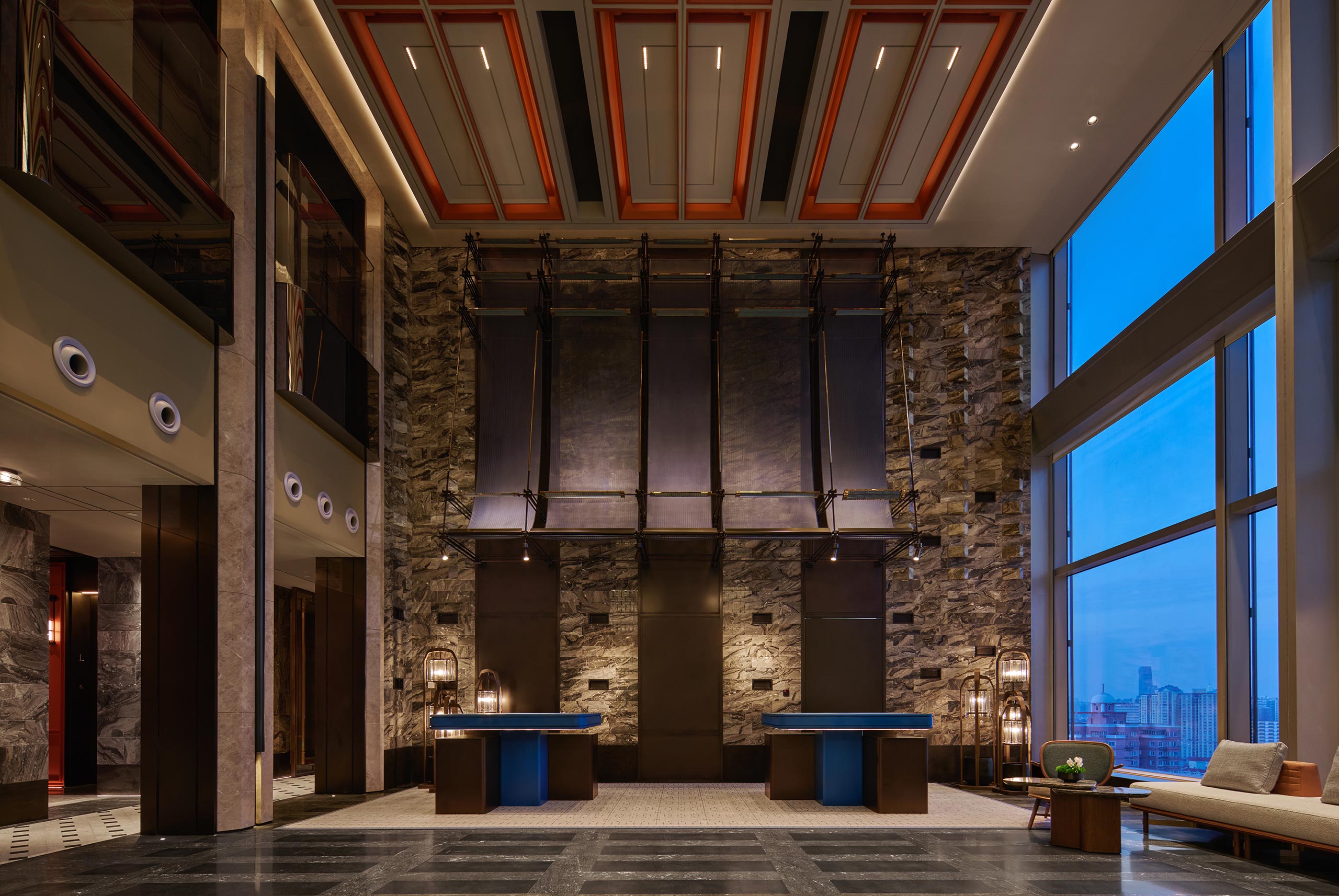 Luxurious hotel lobby with tall stone walls, modern dark fixtures, and large windows showing a cityscape at dusk. Elegant and sophisticated ambiance.