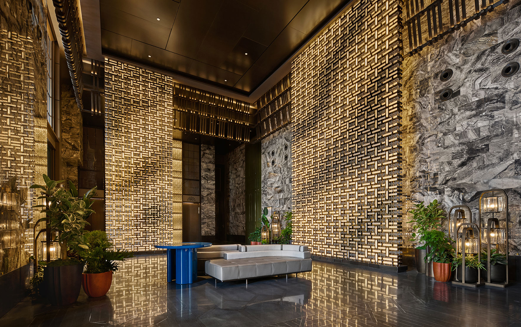 Luxurious modern lobby with illuminated lattice-patterned walls, sleek gray seating, large potted plants, and a vibrant blue table, conveying opulence.