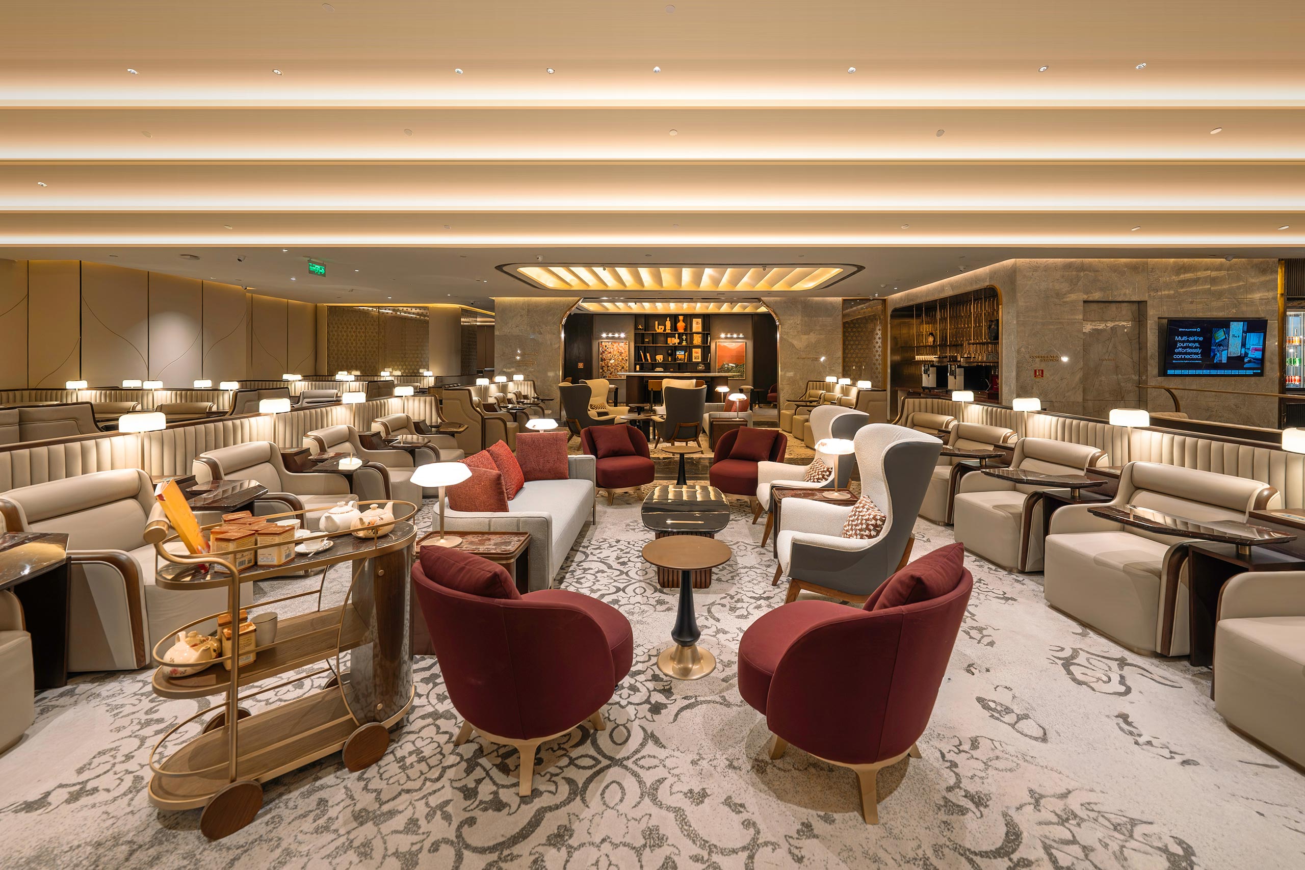Luxurious lounge with plush red and gray chairs, elegant sofas, warm lighting, decorative carpet, and a sophistication bar in the background. Cozy ambiance.