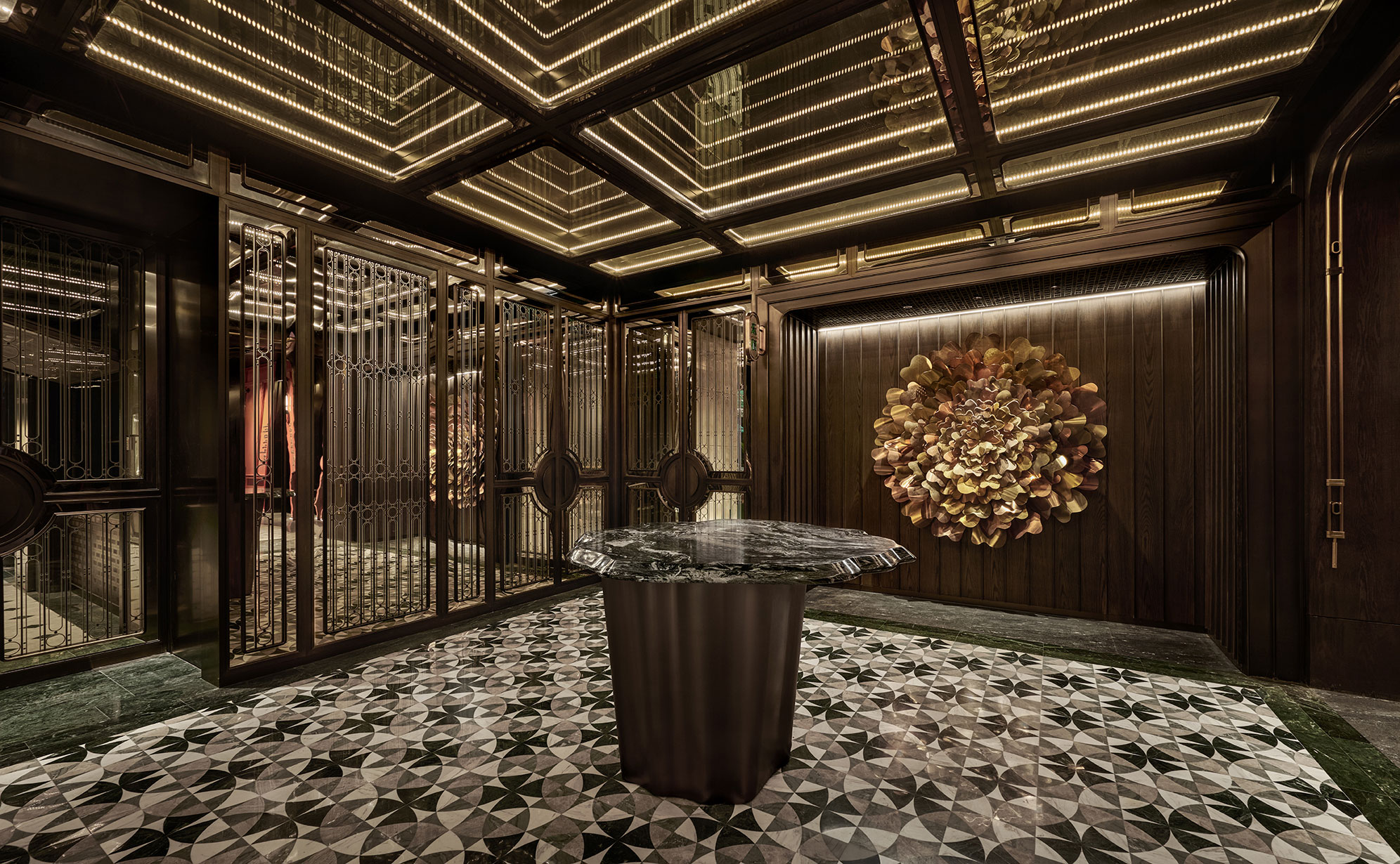 Luxurious lobby with patterned tile flooring, ornate reflective ceiling, and a central marble table. A flower sculpture adorns the dark-paneled wall.