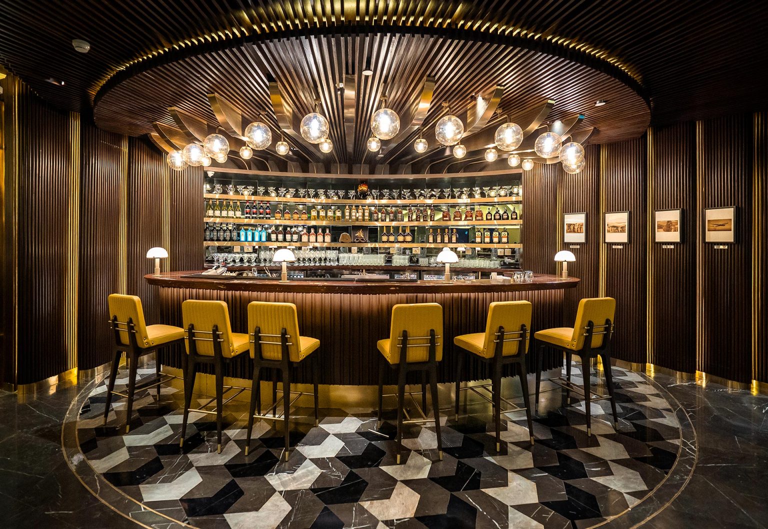 A stylish bar with a semi-circular counter, five yellow chairs, and warm globe lights. Shelves behind hold various bottles, creating a cozy, elegant ambiance.