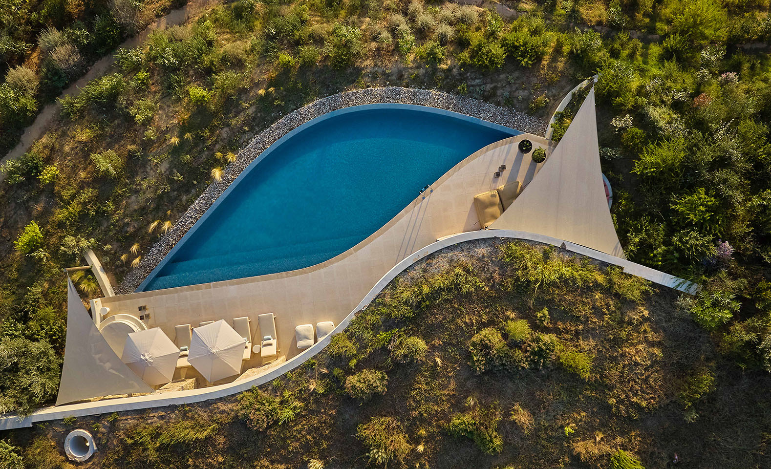 Top-down aerial view of a private leaf-shaped swimming pool at a luxury villa, surrounded by natural green landscaping, a stone sun deck with lounge chairs, and shade sails.