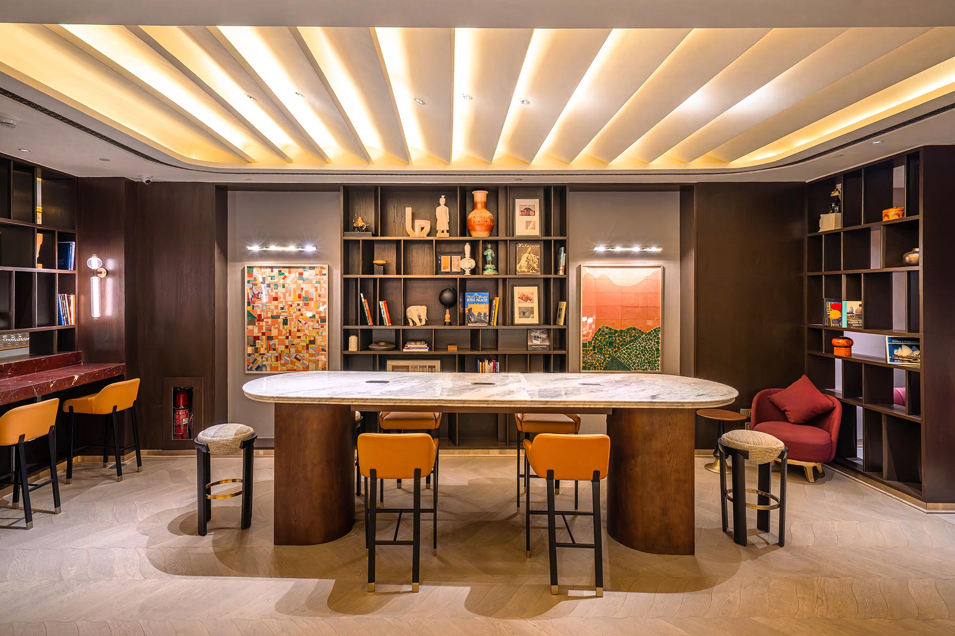 Modern room with an oval marble table, orange chairs, and a backdrop of art-filled dark shelves. Warm lighting creates an inviting, elegant atmosphere.