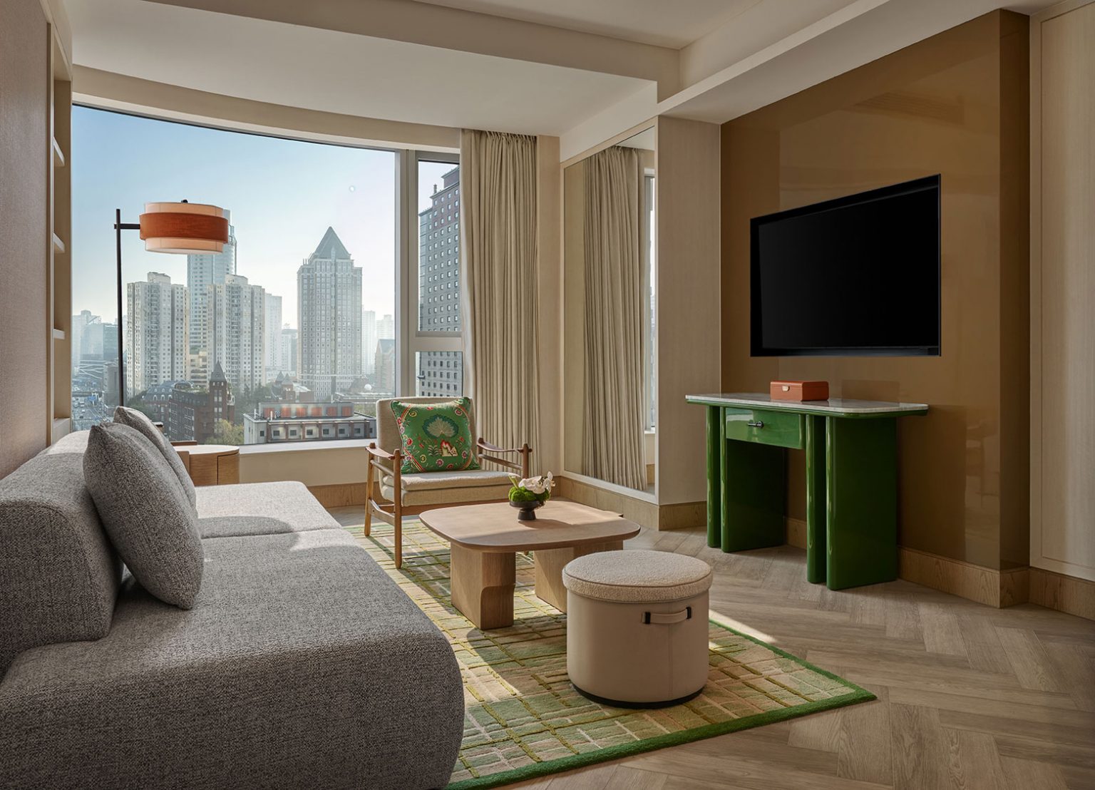 Stylish hotel room with a cityscape view through large windows, featuring a gray sofa, green table, and a flat-screen TV, exuding modern elegance.