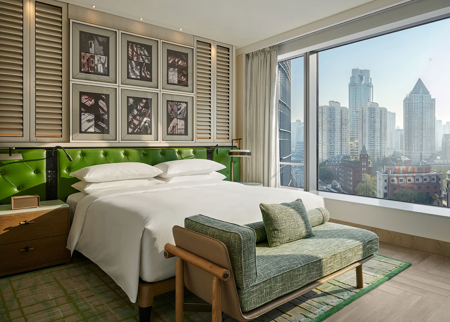 Modern hotel room with a white bed, green headboard, green chaise lounge, and wall art. Large window offers a view of city skyscrapers.