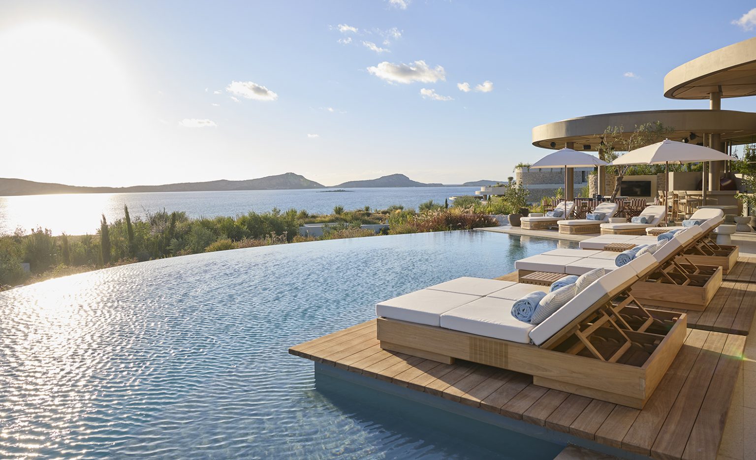 Luxurious infinity pool at sunset with lounge chairs and white umbrellas. Overlooks a serene sea and distant hills, creating a tranquil and relaxing atmosphere.