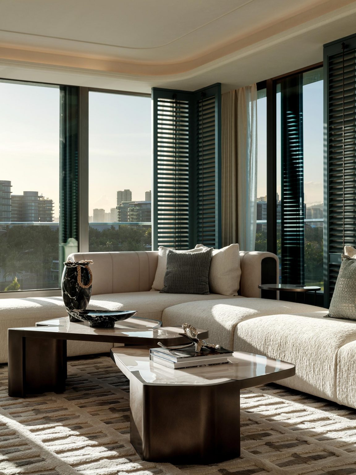 Modern living room with a beige sectional sofa, dark marble coffee table, and city skyline view through large windows with open shutters. Cozy and elegant ambiance.