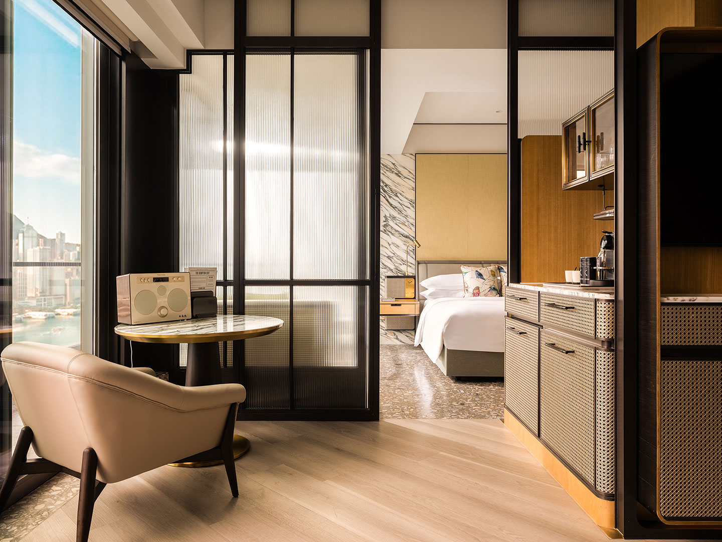 Interior view of a luxury harbour suite at Kimpton Tsim Sha Tsui, featuring a woven-front minibar console in the foreground, a light leather armchair next to a marble table with a retro radio speaker, and a sliding fluted-glass partition opening into the bedroom area.