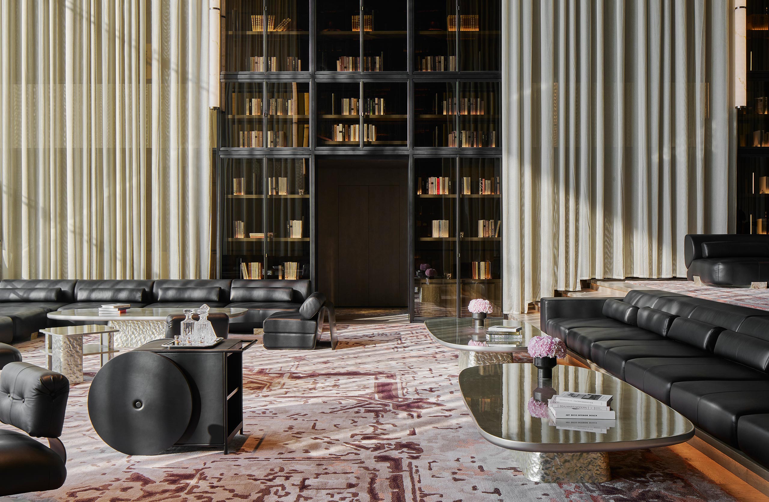 Luxurious lounge with black leather sofas, glass coffee tables, and tall bookshelves. Soft lighting and plush rug create a sophisticated ambiance.