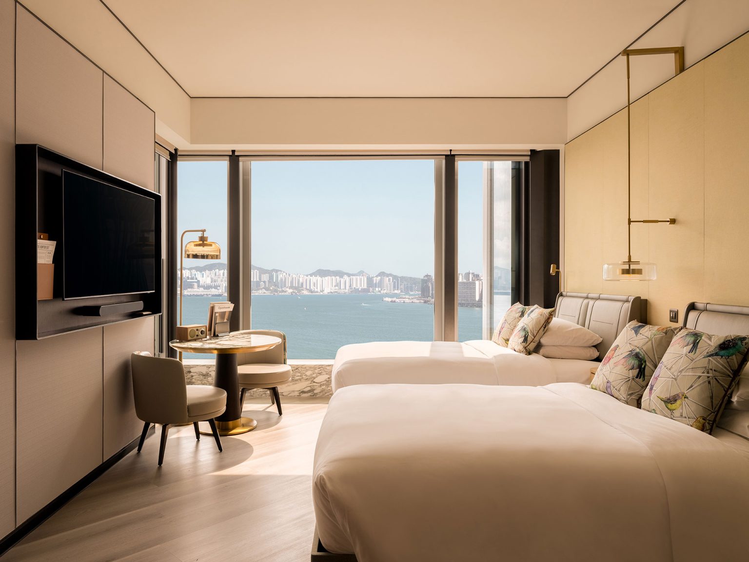 Modern hotel twin guest room at Kimpton Tsim Sha Tsui featuring two beds with patterned pillows, a wall-mounted flat-screen TV, a small dining table with chairs, and a large picture window offering an expansive view of Victoria Harbour and the Hong Kong skyline.