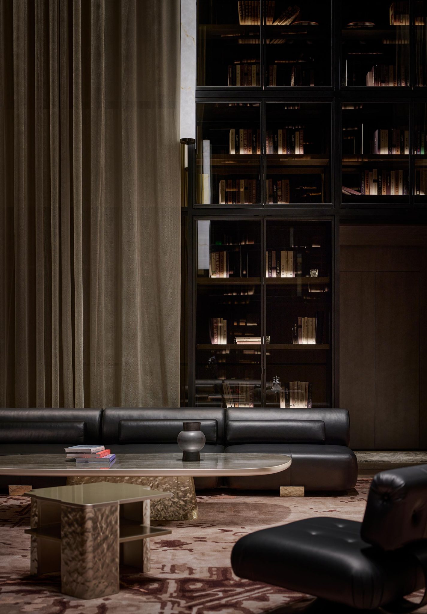 Luxurious living room with dark leather sofa, modern coffee table, ornate shelves with books, floor-to-ceiling curtains, and a plush rug, creating a warm ambiance.