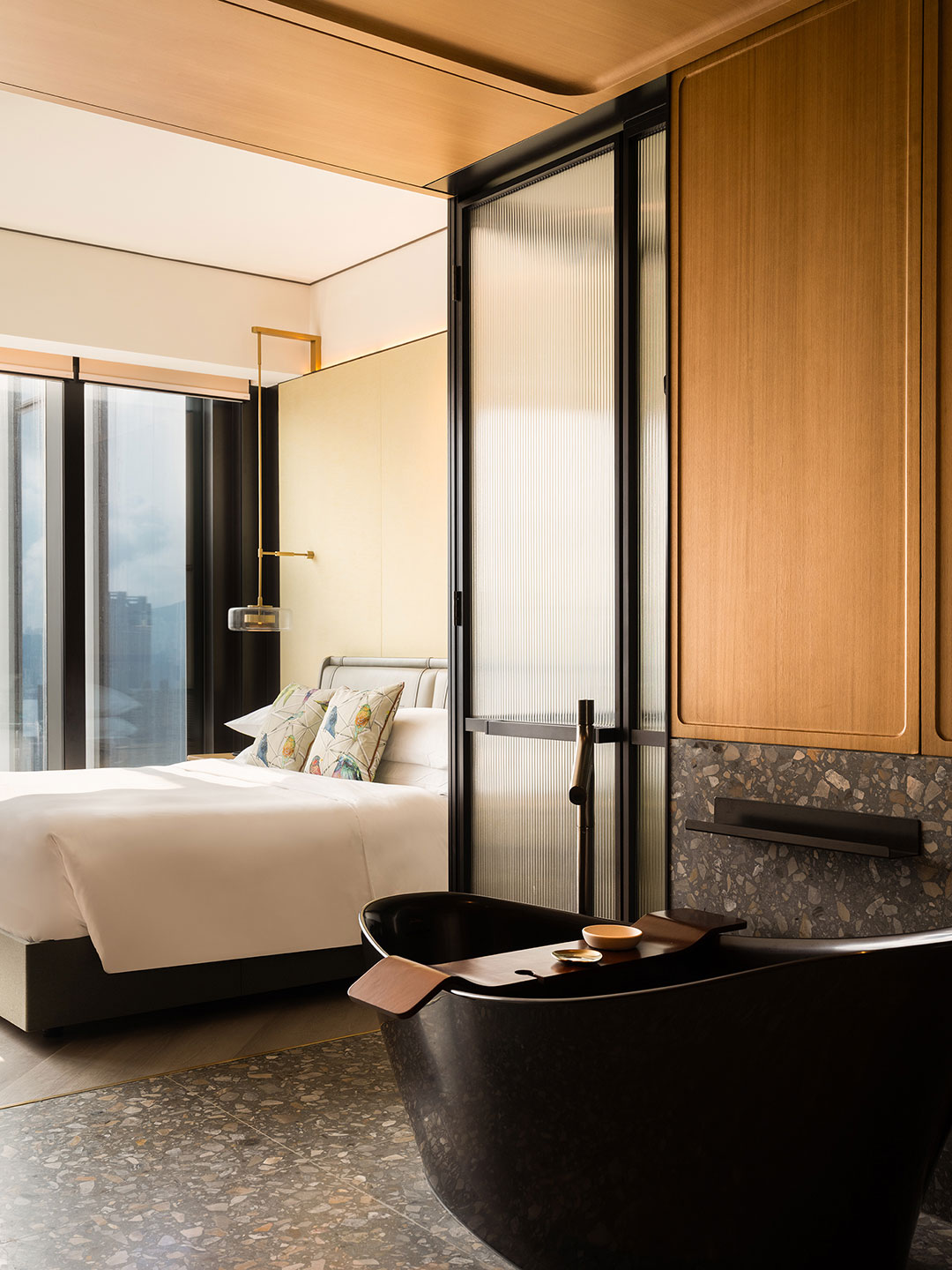 Luxury hotel room interior showcasing a sleek, freestanding black bathtub on a terrazzo floor in the foreground, partially separated from a plush king bed and large windows by a textured glass sliding door.