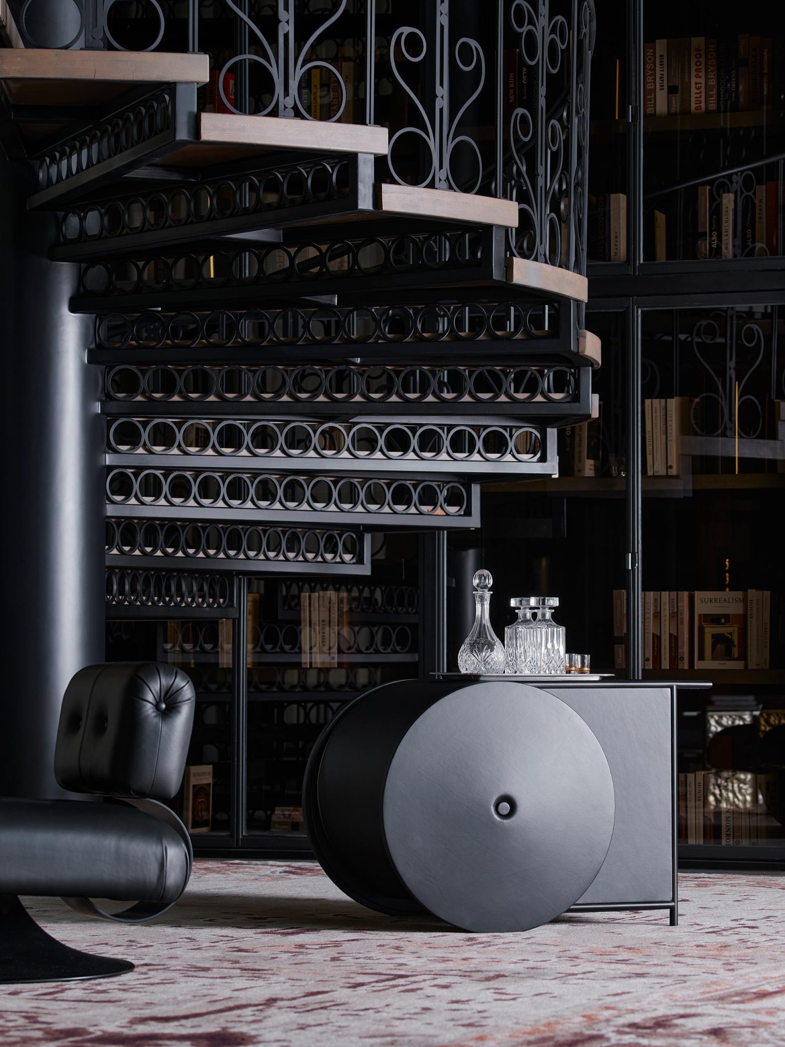 Elegant spiral staircase with intricate wrought iron design, flanked by bookshelves. A plush black leather chair and glass decanters on a modern table below.