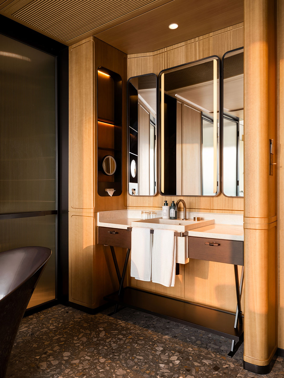 Chic hotel bathroom vanity featuring a double mirror setup with integrated LED lighting, a white undermount sink, warm wood paneling, and an elegant dark terrazzo tile floor.