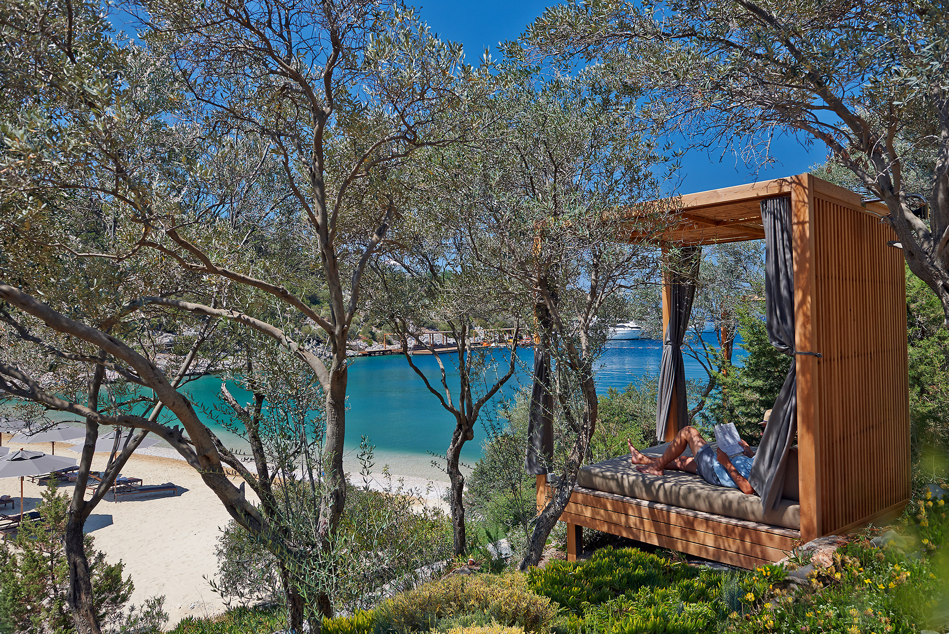 Private wooden luxury cabana nestled among lush olive trees on a hillside, overlooking a sandy beach cove and a vibrant turquoise sea.
