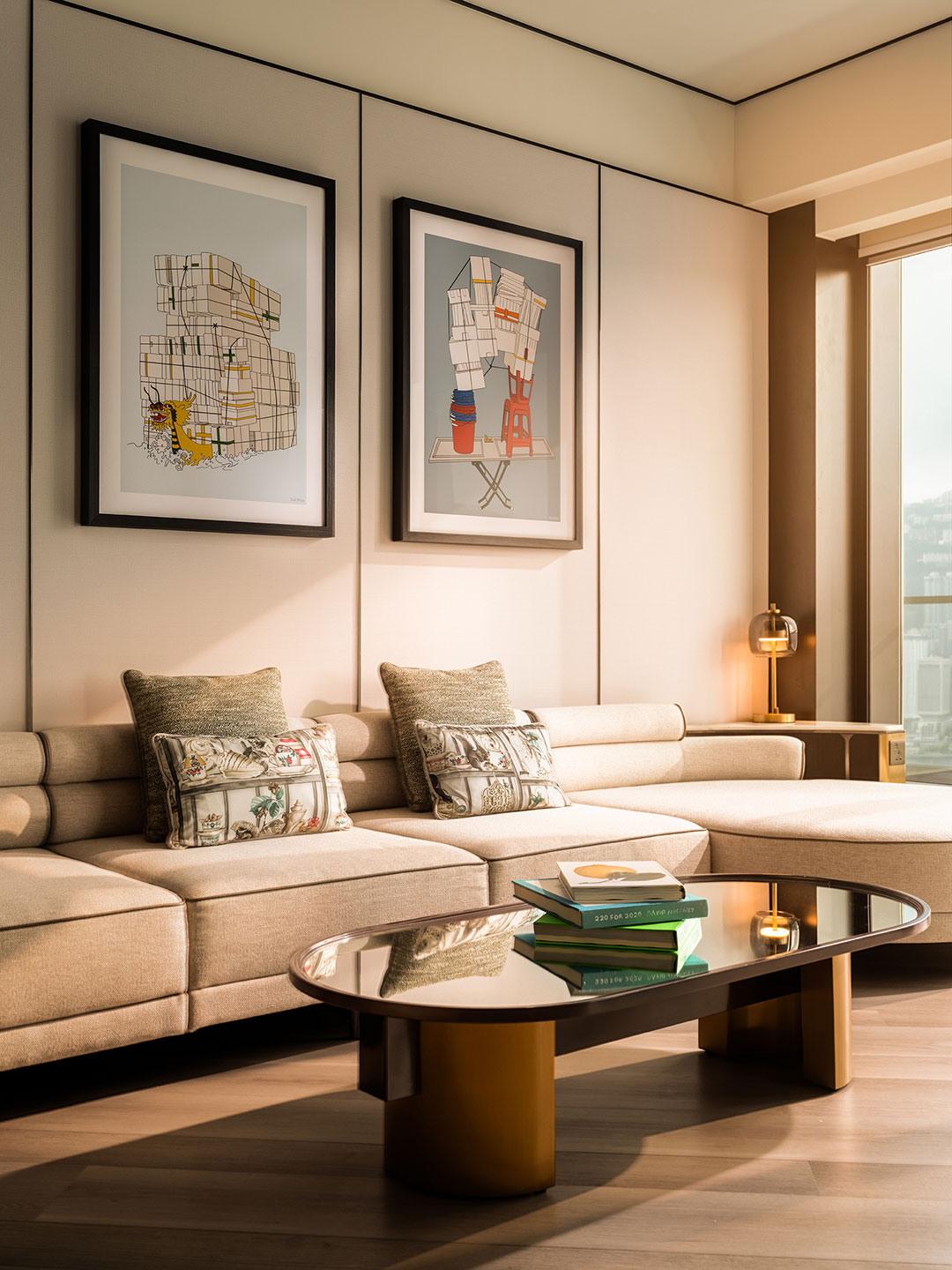 Cozy living room area of a hotel suite at Kimpton Tsim Sha Tsui, showcasing a beige sectional sofa with decorative cushions, framed graphic artwork on the wall, and a mirrored coffee table stacked with books.