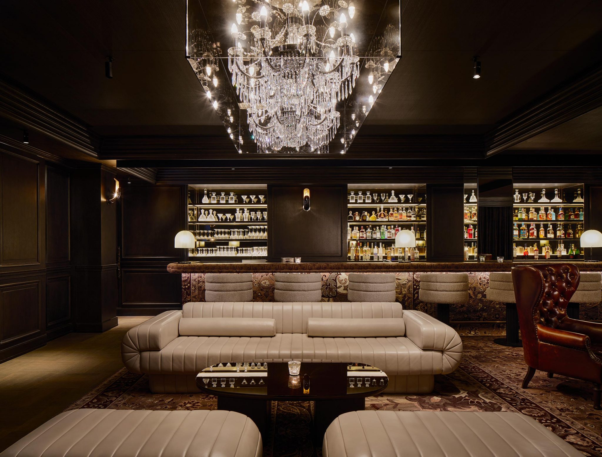 Luxurious bar interior with plush beige couches, a grand crystal chandelier, and shelves filled with bottles, creating an elegant and sophisticated atmosphere.
