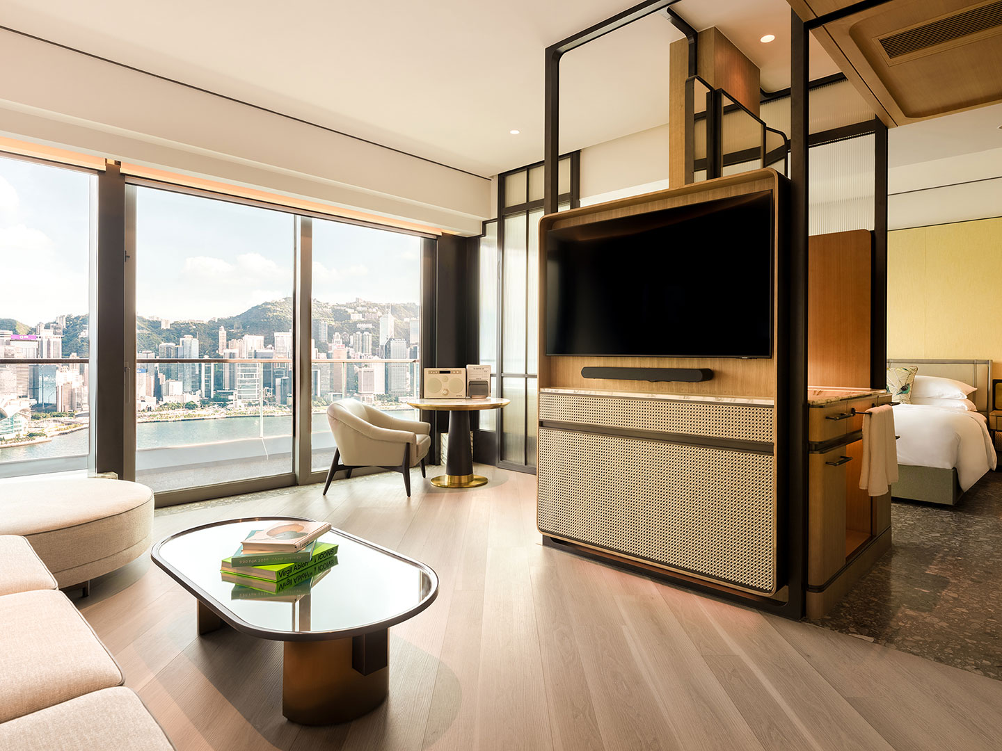 Spacious luxury harbour suite layout showing the open-concept living area with a glass coffee table, a rotating flat-screen TV partition wall, and large floor-to-ceiling windows overlooking Victoria Harbour and Hong Kong island skyscrapers.