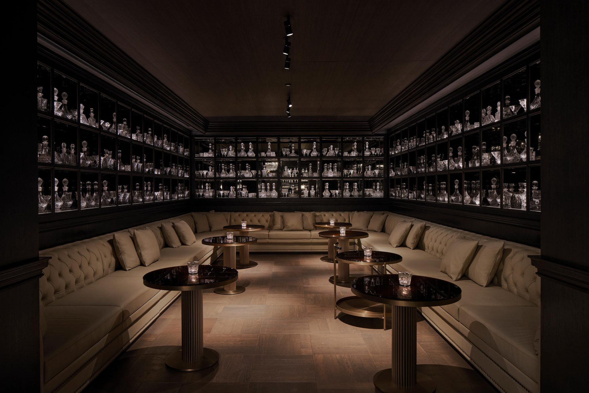 Dimly lit lounge with beige tufted sofas, round tables, and shelves lining the walls filled with glass decanters and bottles.