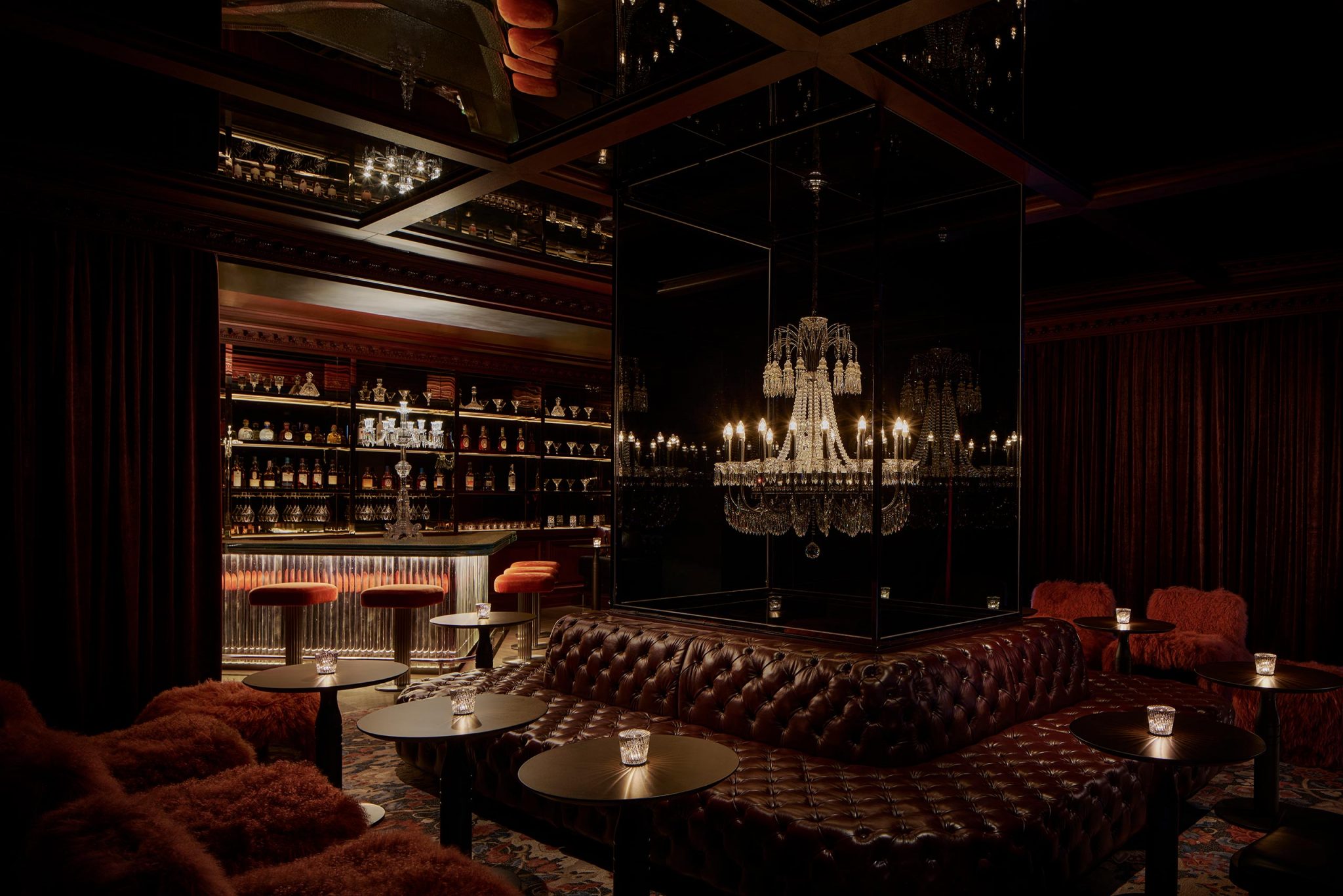 Dimly lit lounge with brown tufted leather seating, small round tables, red barstools, a mirrored chandelier, and a stocked bar in the background.