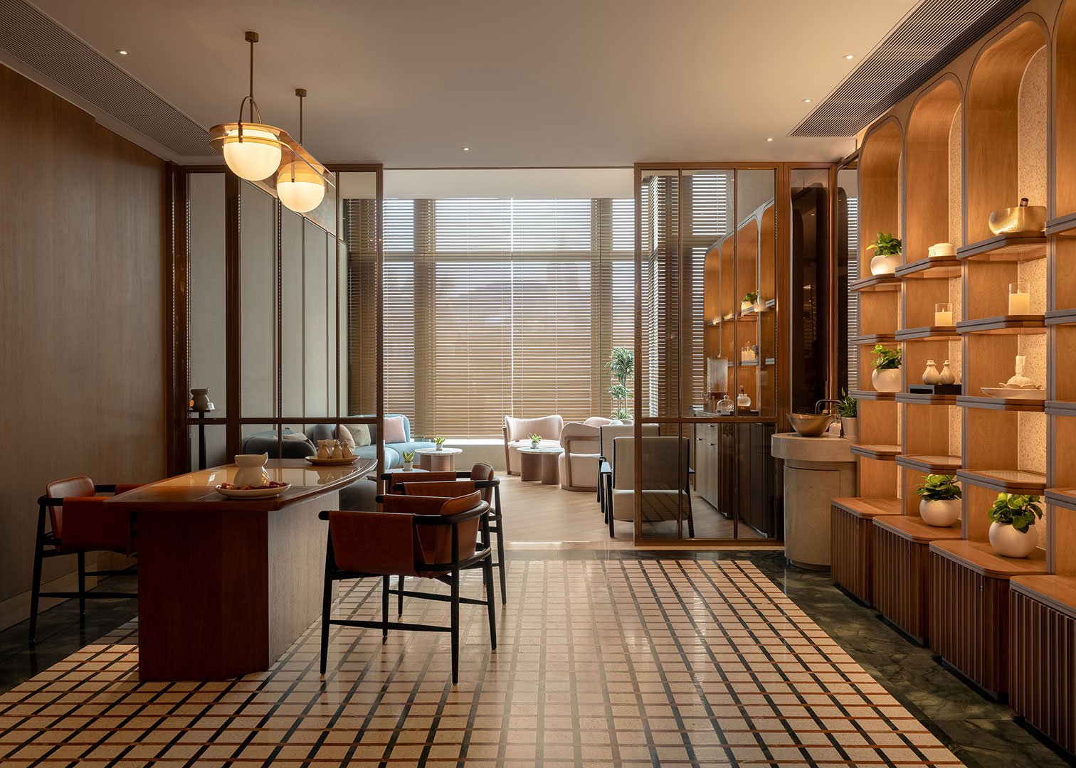 The elegant reception and lounge area of the spa at Kimpton Tsim Sha Tsui, featuring a geometric patterned tile floor, a wooden consultation desk with leather chairs, warm hanging globe pendant lights, and built-in wooden arched display shelving holding small plants and wellness products.