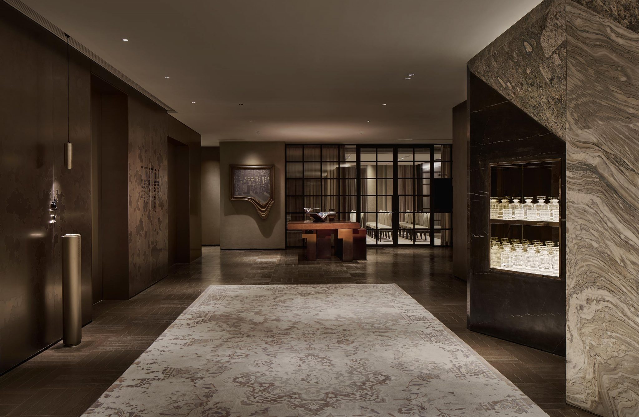 Elegant hotel lobby with dim lighting, featuring a plush, patterned carpet leading to a wooden counter flanked by decorative shelving and art.