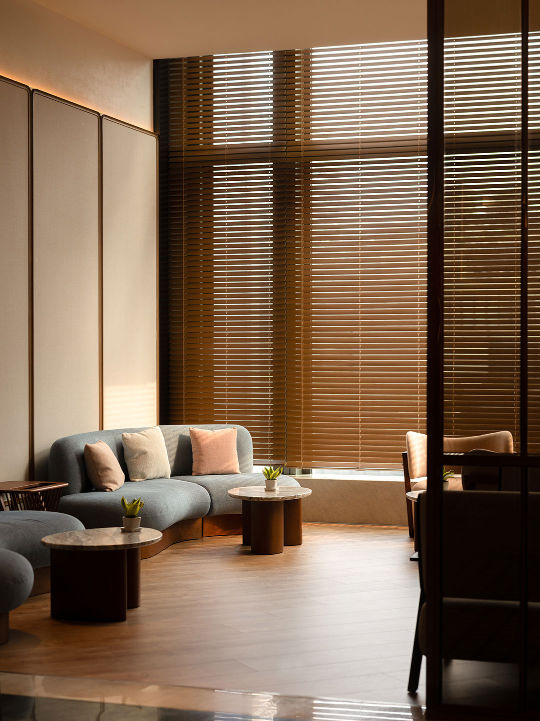 A serene spa relaxation lounge bathed in warm light, showcasing a curved light blue sofa with plush accent pillows, small round marble-topped coffee tables with potted succulents, and large floor-to-ceiling windows covered with horizontal wooden blinds.