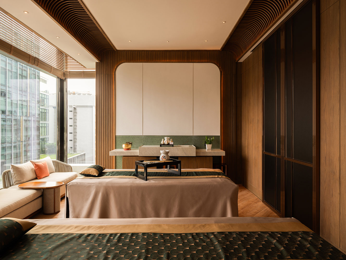 A luxurious couples massage room inside the hotel spa, featuring two parallel treatment tables dressed in neutral linens, a cozy window-side seating banquette, fluted wood wall panels, a custom marble wash station, and a textured geometric ceiling detail.
