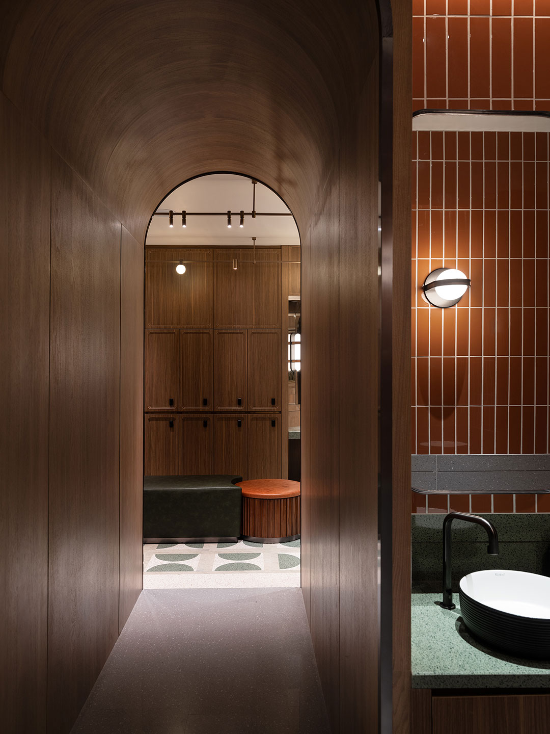 A sophisticated spa changing room area viewed through a wood-paneled archway, highlighting a green terrazzo vanity countertop with a modern black-rimmed vessel sink, gloss orange-brown subway wall tiles, and wooden lockers with a seating bench in the background.