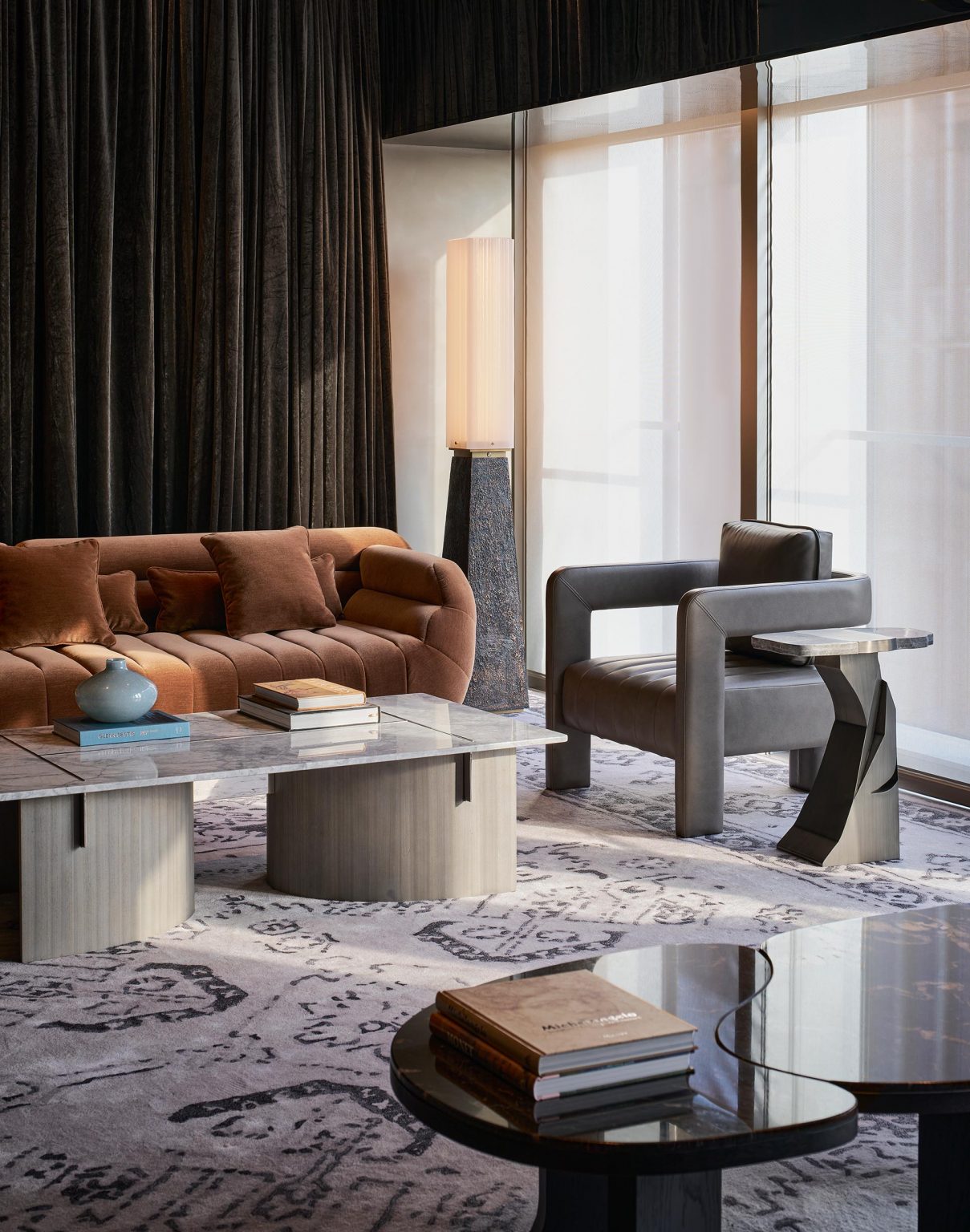 Modern living room with a brown sofa, gray armchair, marble coffee tables, books, a blue vase, patterned rug, large windows with sheer curtains, and a tall floor lamp.