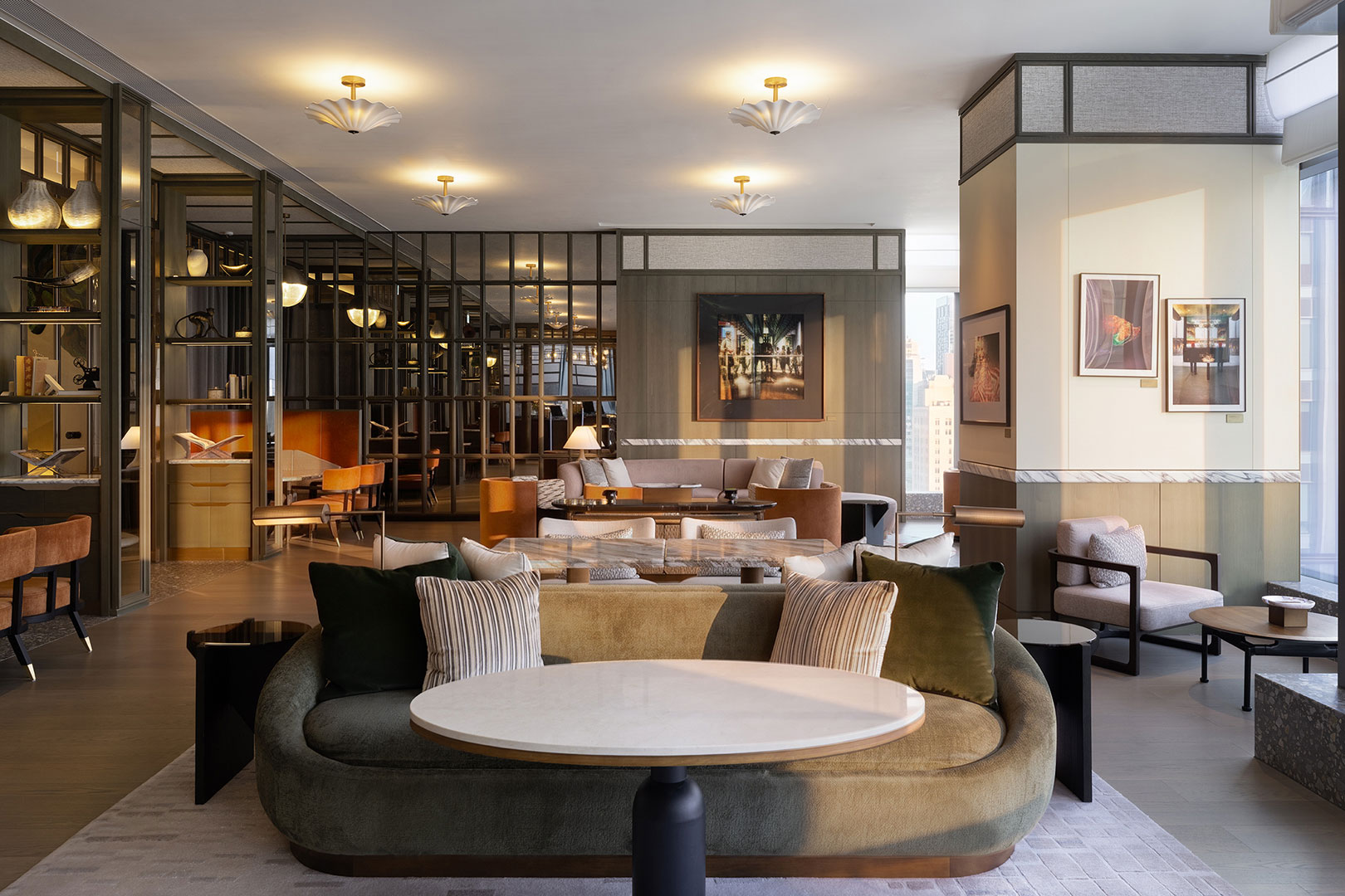 The stylish executive lounge at Kimpton Tsim Sha Tsui, showcasing a curved velvet sofa in the foreground with a round marble table, multiple intimate seating groups, decorative glass and metal partitions, and curated artwork on the walls under warm scalloped ceiling lights.