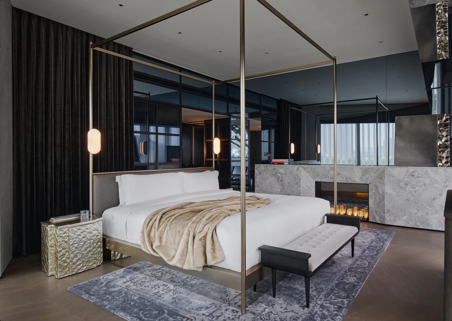 Luxurious hotel bedroom with a four-poster bed, soft beige blanket, sleek black and gold accents, elegant fireplace, and modern decor. Cozy ambiance.