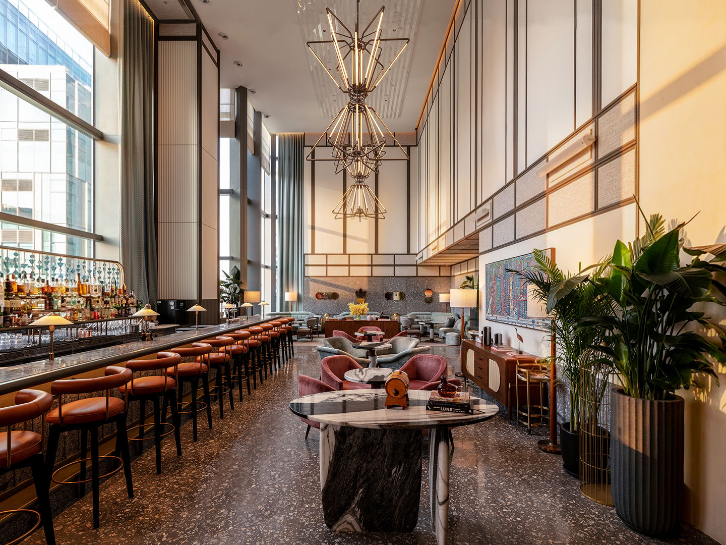 The dramatic, high-ceilinged Birdsong dining and bar area featuring a long marble counter bar with tan leather barstools, striking multi-tiered geometric wire chandeliers, terrazzo flooring, velvet armchairs around marble tables, and expansive windows framing the city view.