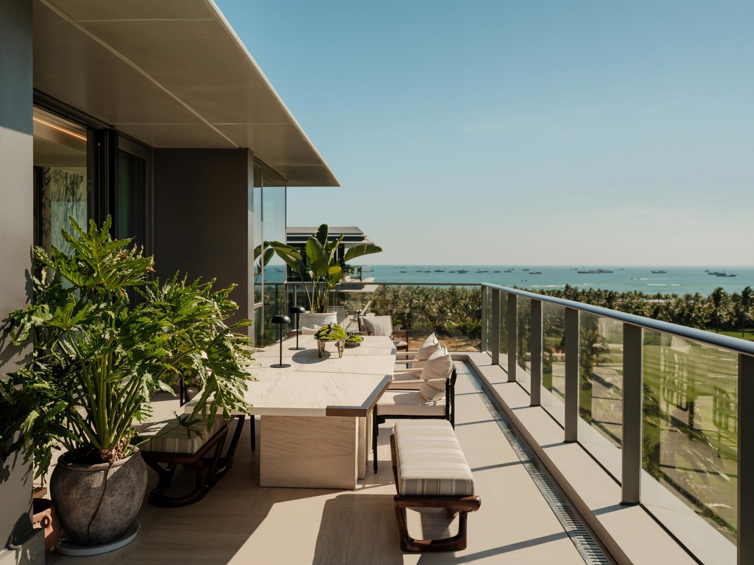 Modern rooftop terrace with lush potted plants, a sleek dining table, and glass railings overlooking a scenic ocean view under a clear blue sky.