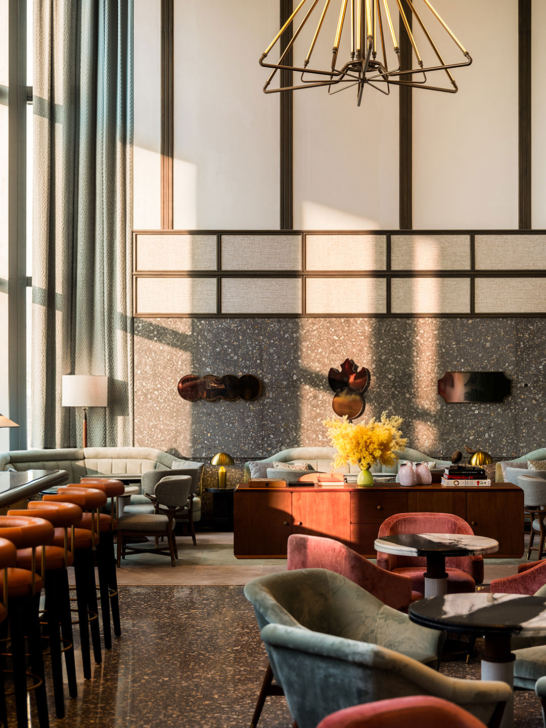 A detailed view of the Birdsong dining room lounge area, highlighting low-slung velvet chairs in sage green and dusty rose, a wooden credenza topped with a bright yellow floral arrangement, a grey terrazzo accent wall with decorative mirrors, and warm sunlight streaming through high windows.