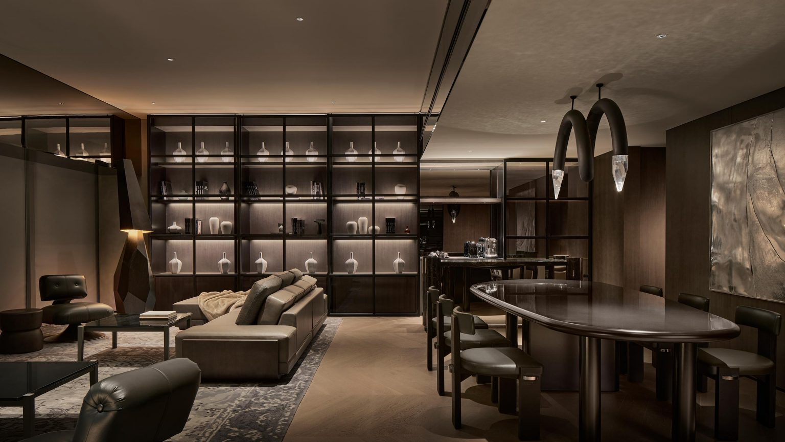 Luxury lounge with dim lighting, featuring plush seating, a long dining table, and a backlit shelf displaying elegant decor. Modern, sophisticated ambiance.
