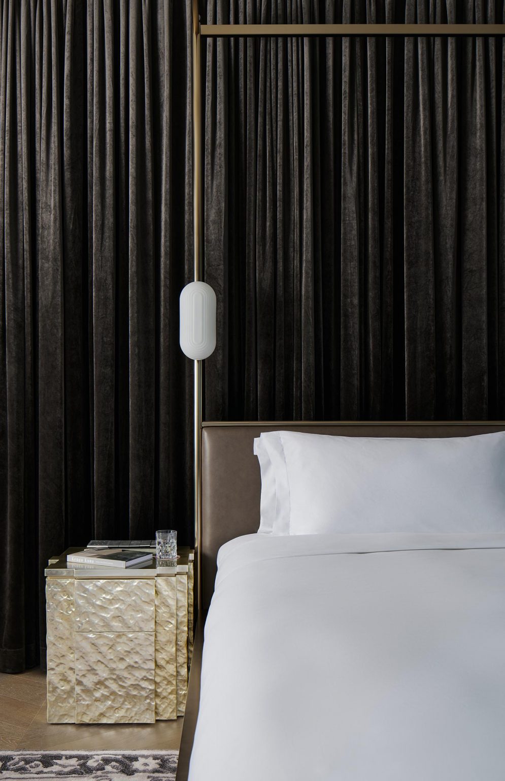 Luxurious bedroom with white bedding, dark drapes, and a glowing wall lamp beside a textured, gold nightstand holding books and a glass. Peaceful ambience.