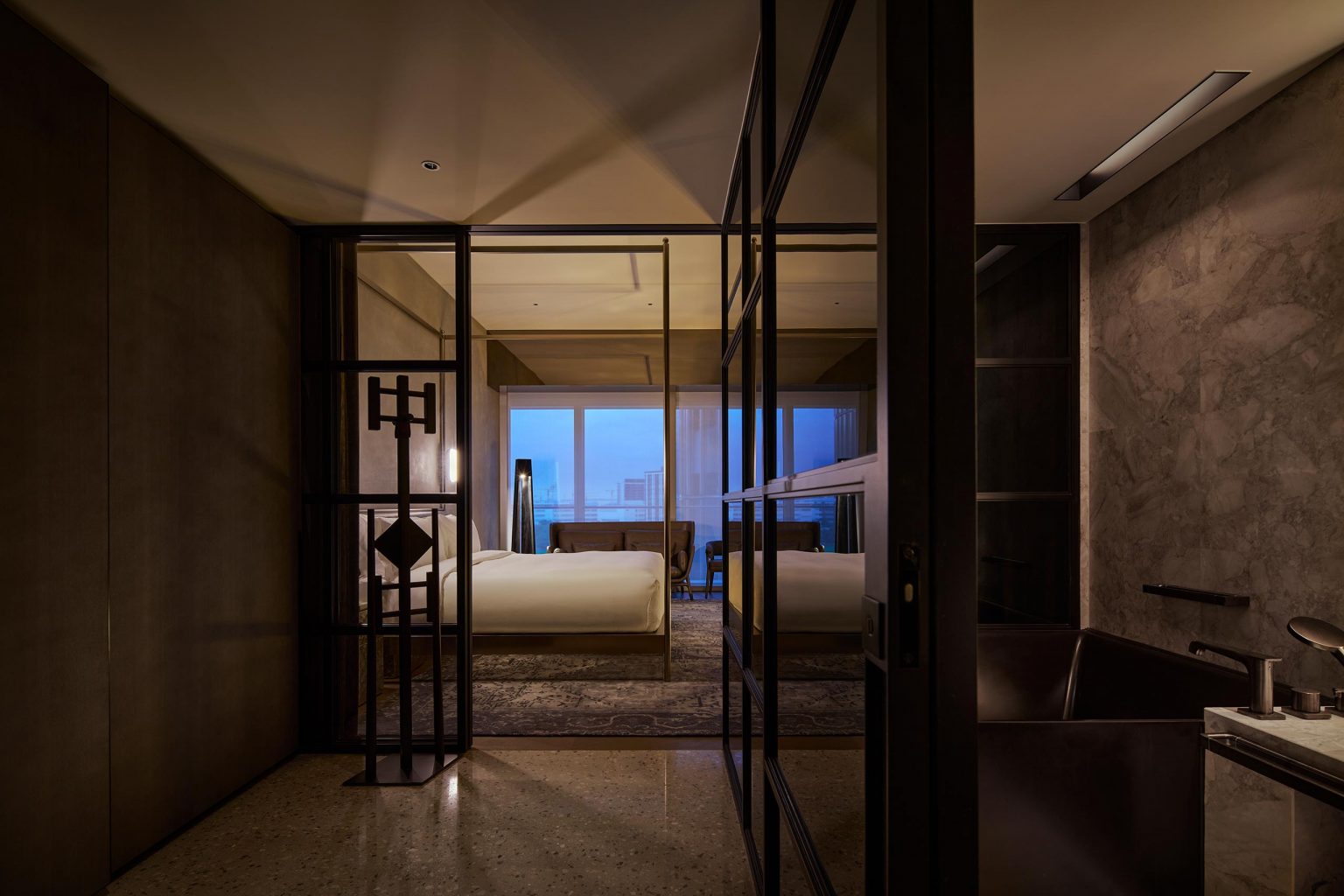 Luxurious hotel room with moody lighting, featuring a large bed, floor-to-ceiling windows with city view, and elegant décor. A modern bathroom is visible.