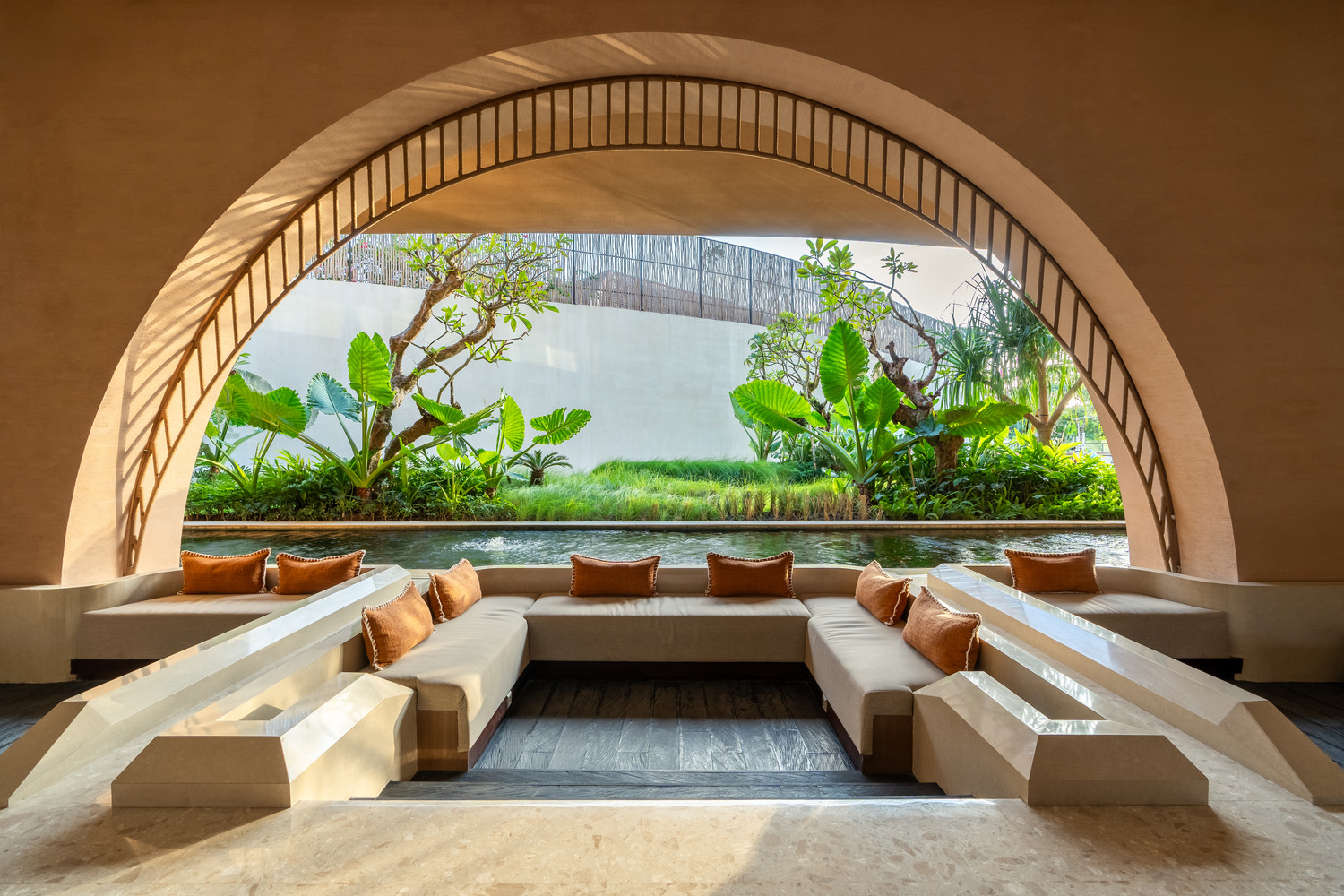 Elegant lounge area with white sofas and brown cushions under a large archway. Lush greenery and a serene water feature visible outside. Peaceful ambiance.